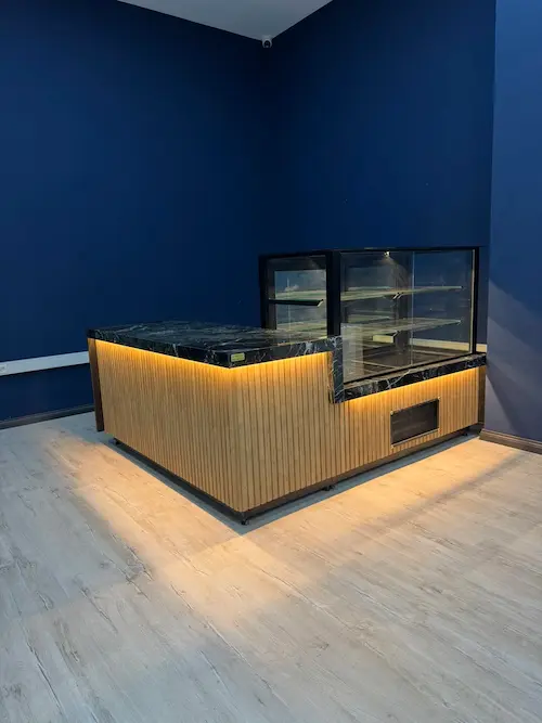 Açık renk ahşap dikey oluklu panelli, siyah mermer tablalı, LED aydınlatmalı köşe cafe kasa bankosu ve entegre camlı pasta dolabı