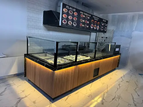 Meşe rengi ince dikey çıtalı panelli, siyah mermer üst tablalı, cam siperlikli ve LED aydınlatmalı yemek servis tezgahı ve entegre kasa bankosu