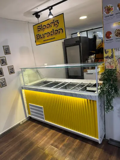 Canlı sarı dikey panelli ön dekorlu, beyaz mermer üst tablalı, düz cam korumalı modern fast food sipariş ve servis bankosu