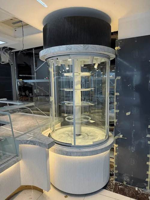 Beyaz oluklu panelli, mermer detaylı, kendi etrafında dönen raflı, silindir formunda lüks pasta ve tatlı teşhir kulesi